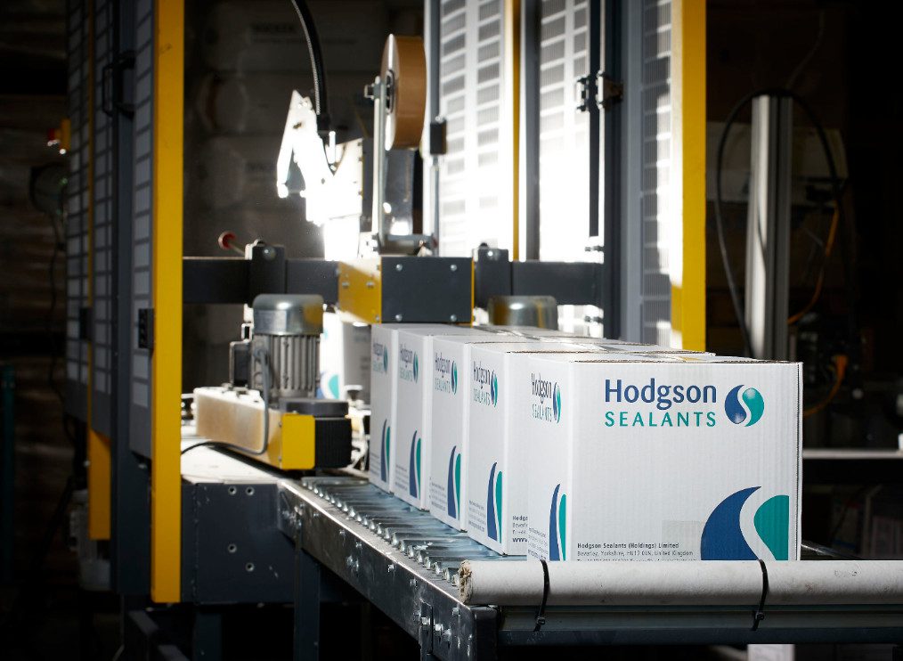 A set of Hodgson Sealant products packed in a box on a conveyor belt.